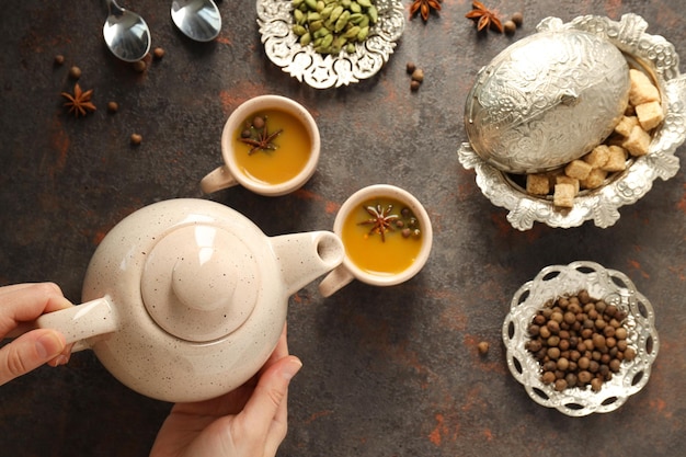 Masala tea in cups spices and teapot in female hands on wooden background top view