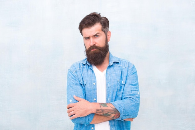 Masculinity and male beauty concept Well groomed macho Brutal handsome hipster man on grey wall background Bearded man trendy hipster style Hipster with beard and mustache wear denim shirt