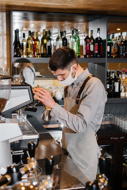 A masked barista prepares delicious coffee at the bar in a cafe. The work of restaurants and cafes during the pandemic.