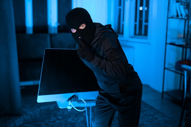 Masked thief carrying tv or desktop screen in dark apartment