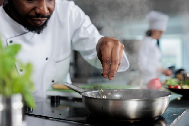 Master chef wearing cooking uniform garnishing gourmet dish with fresh chopped herbs in restaurant kitchen. Gastronomy expert using pan to cook delicious food for dinner service.