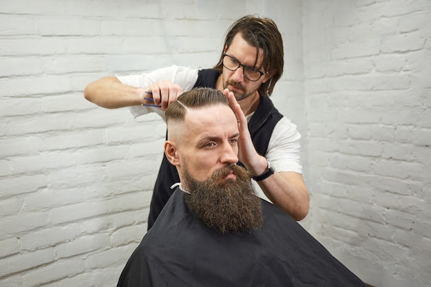 Master cuts hair and beard of men in the barbershop, hairdresser makes hairstyle for a young man