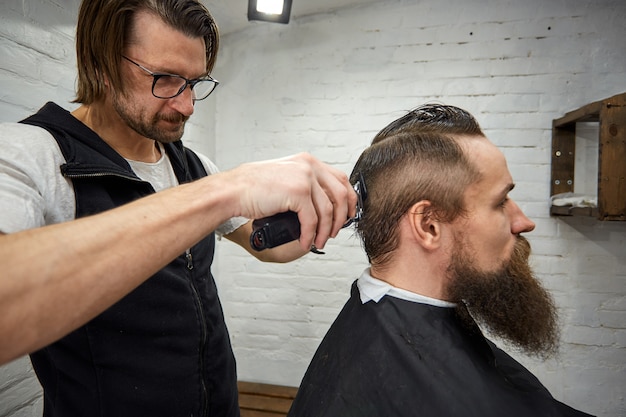 Master cuts hair and beard of men in the barbershop, hairdresser makes hairstyle for a young man