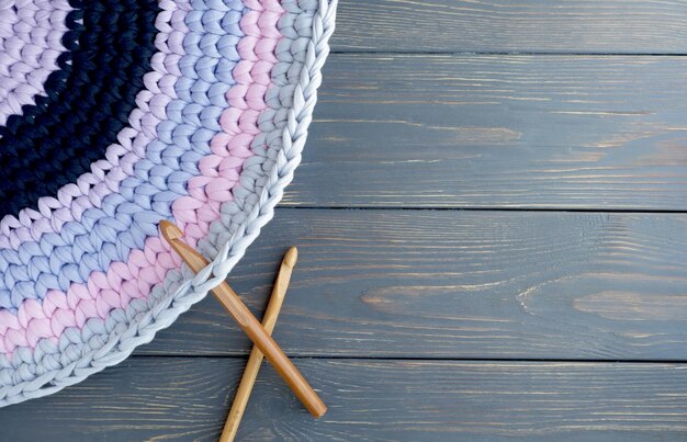 A mat made of knitted yarn and a crochet hook on a gray wooden background with space for text Home needlework