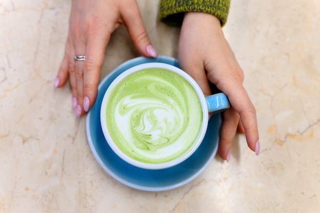 Matcha green tea latte with a pattern of milk foam in a blue ceramic cup and women's hands on the table