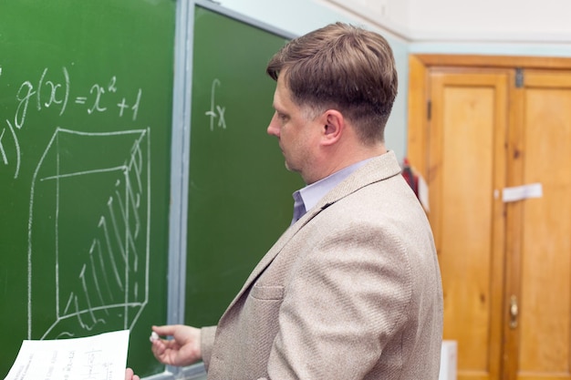 Mathematics teacher writes in chalk on a blackboard with his back to the class