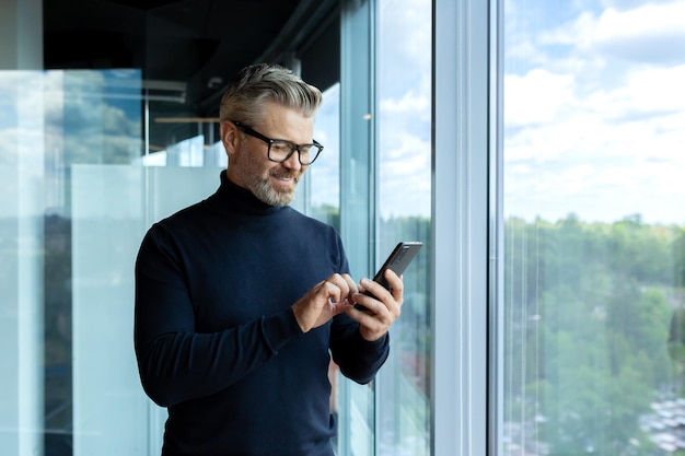 Mature adult man inside office standing near window businessman typing message on phone and browsing