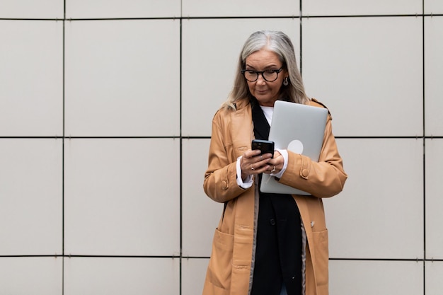 Mature adult woman freelancer with gray hair in glasses standing on the street with a laptop