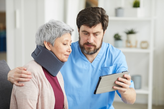 Mature bearded doctor showing something on tablet pc to his senior patient who has a neck injury