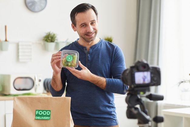 mature bearded man holding organic salad box while recording food delivery service review in kitchen interior