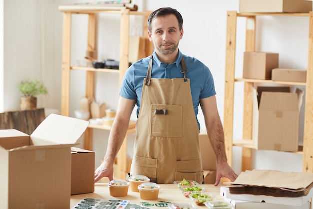 mature bearded man wearing apron and smiling standing by wooden table with individual food portions ready for packaging, worker in food delivery service