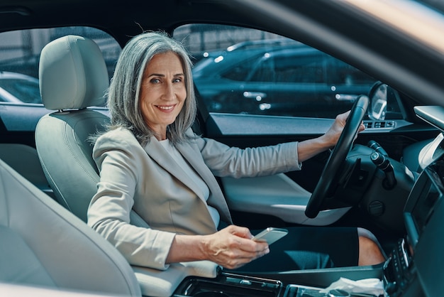 Mature beautiful woman in smart casual wear looking at camera and smiling while driving car