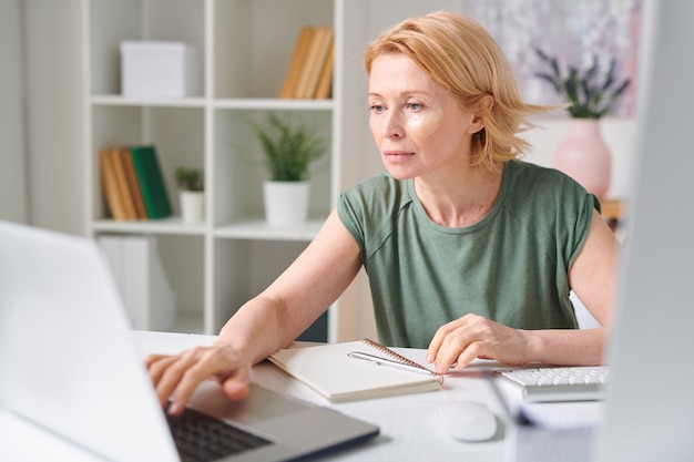 Mature blond casual female with undereye patches looking at laptop display while working remotely at home during self isolation