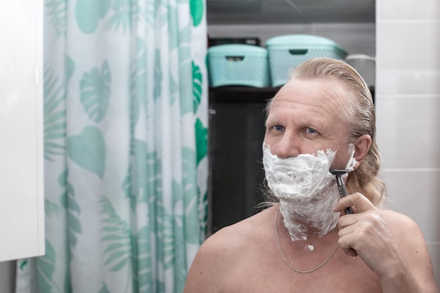 A mature blond man with plenty of foam on his face shaves with a safety razor in the bathroom Men39s hygiene and facial care