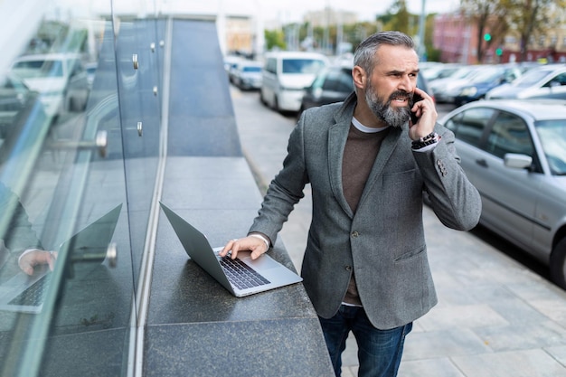 Mature business man in a jacket with a laptop talking on the phone outside