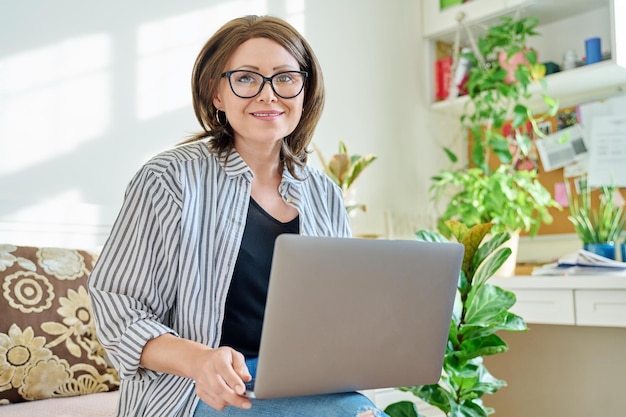 Mature business woman working on couch using laptop looking at camera