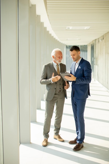 Mature businessman and his young colleague discussing finantial report on digital tablet in the office corridor