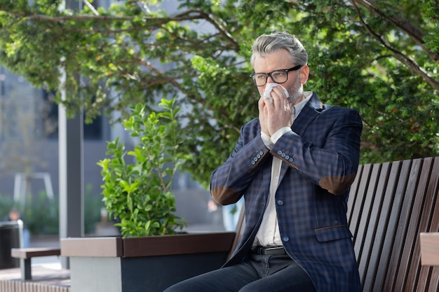 Mature businessman working outdoors with laptop senior man sneezing from allergies sitting on park