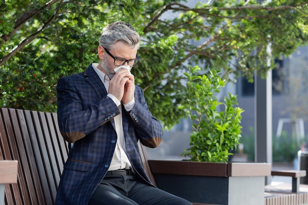 Mature businessman working outdoors with laptop senior man sneezing from allergies sitting on park