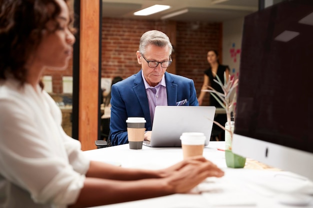 Mature Businessman Working At Table On Laptop In Open Plan Office With Colleagues In Background