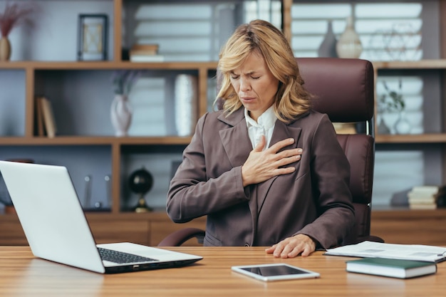 Mature businesswoman in home office feeling unwell hand on chest with laptop and tablet on her desk