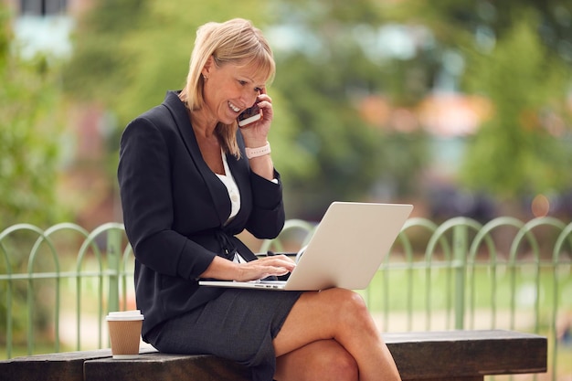 Mature Businesswoman Sitting On City Park Bench Working On Laptop And Talking On Mobile Phone