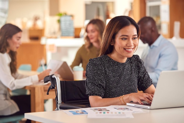 Mature Businesswoman In Wheelchair Sitting At Desk Working On Laptop In Busy Office