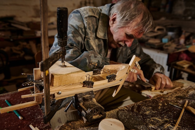 Photo a mature carpenter works on a wooden toy sailboat in his workshop. craftsman makes handmade wooden objects in his workshop