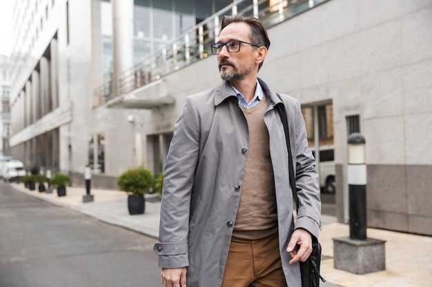 mature caucasian businessman in eyeglasses looking aside while standing near office building at city street