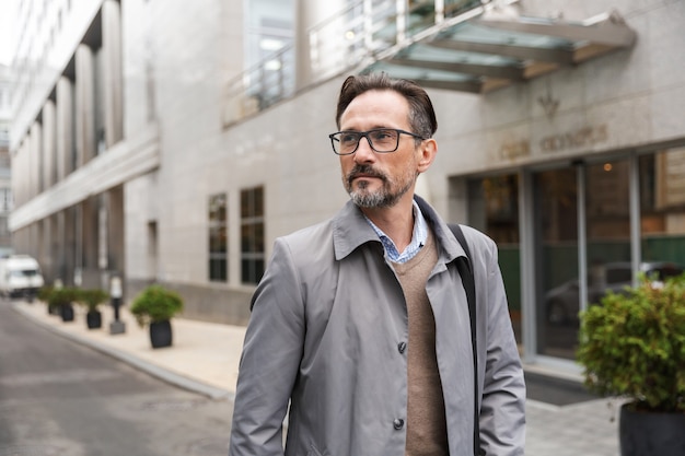 mature caucasian businessman in eyeglasses looking aside while standing near office building at city street