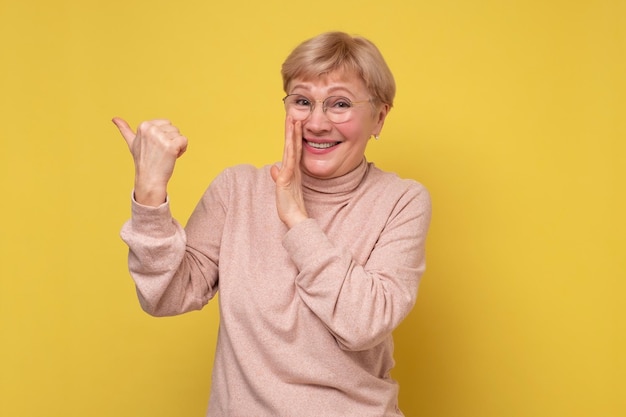 Mature caucasian woman pointing with index finger aside whispering secret behind her hand