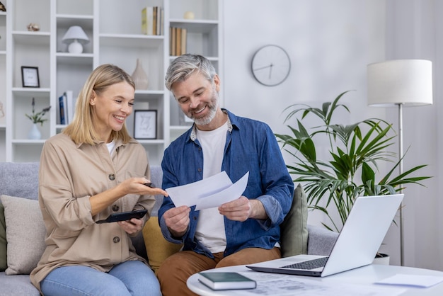 A mature couple in a cozy living room sorts and discusses documents with a laptop and calculator