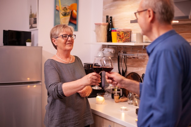 Mature couple looking at each other holding glasses of red wine in the evening. Aged couple in love talking having pleasant conversation during healthy meal.