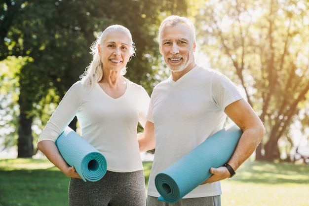 Mature couple standing with yoga carpet in hands in morning park before workout with look at camera