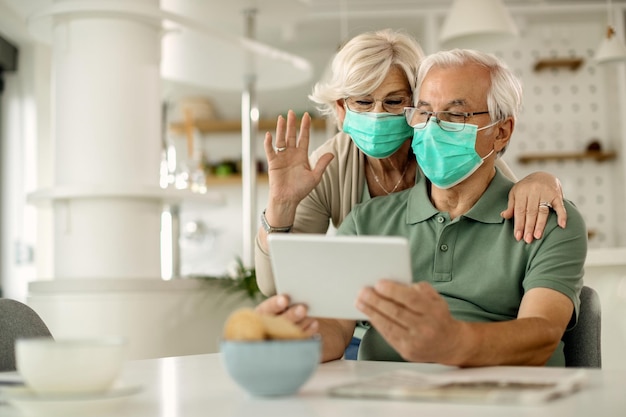 Mature couple with face masks making video call over touchpad at home