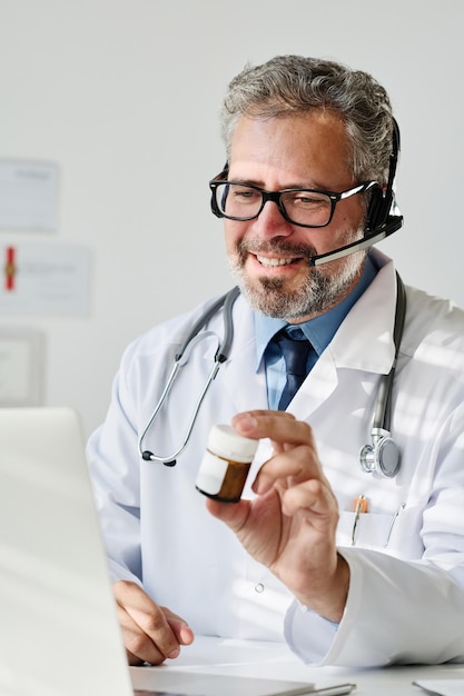 Mature doctor in eyeglasses and headphones prescribing medicine to patient during video call on lapt