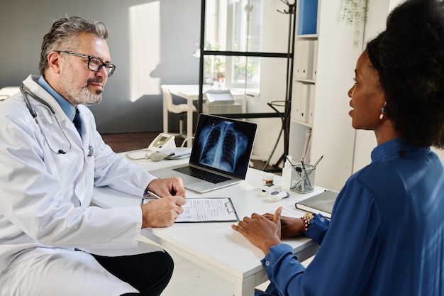 Mature doctor in white coat listening to female patient and making notes in medical card at table