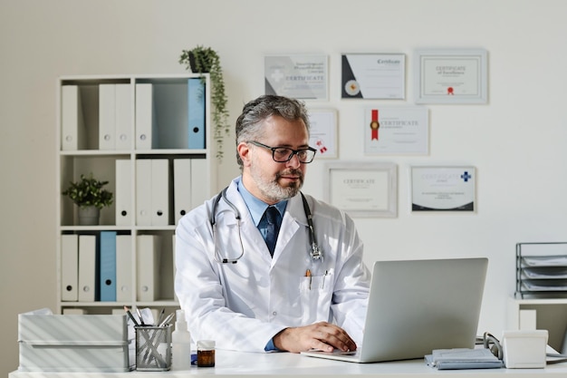 Mature doctor in white coat sitting at table and typing on laptop he working online at office