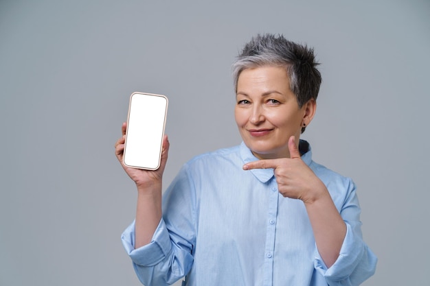 Mature grey hair woman smiling holding smartphone pointing on white screen working or shopping online social media Pretty woman in blue shirt isolated on white background