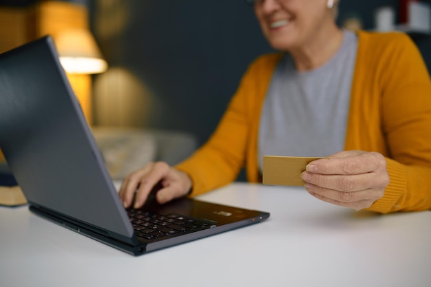 Mature grey-haired woman using bank credit card for online payment on laptop from home office