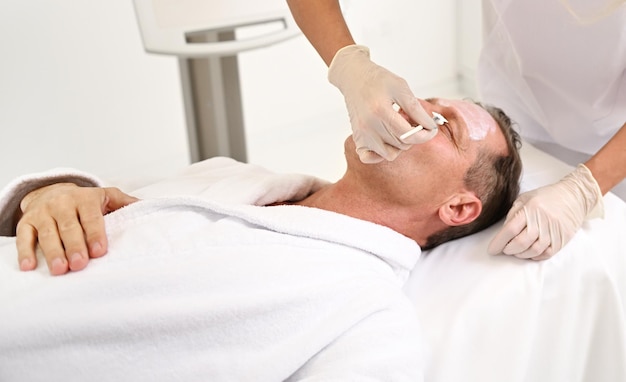 Mature handsome man lying down on a massage table at male beauty treatment for anti-aging and rejuvenation. Beautician applying facial cosmetic on the face of a wellness spa center client