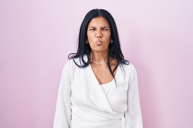 Mature hispanic woman standing over pink background making fish face with lips, crazy and comical gesture. funny expression.