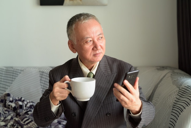 Mature Japanese businessman thinking while holding phone and coffee at home