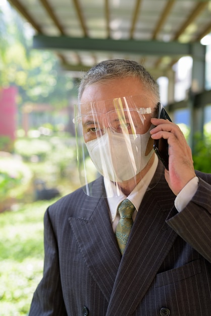 Mature Japanese businessman with mask and face shield talking on the phone at the park