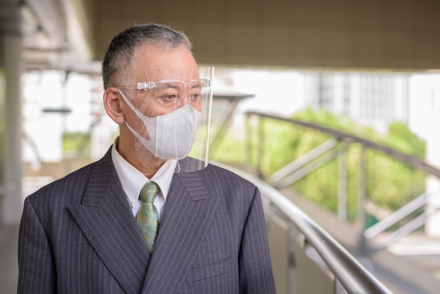 Mature Japanese businessman with mask and face shield thinking at the footbridge