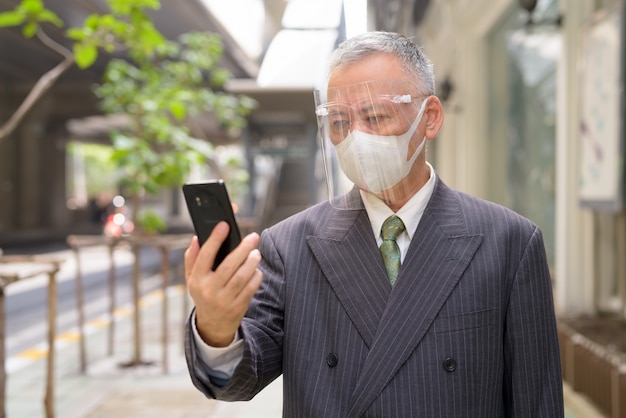 Mature Japanese businessman with mask and face shield using phone in the city