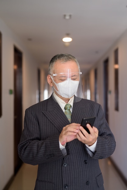 Mature Japanese businessman with mask and face shield using phone in the corridor