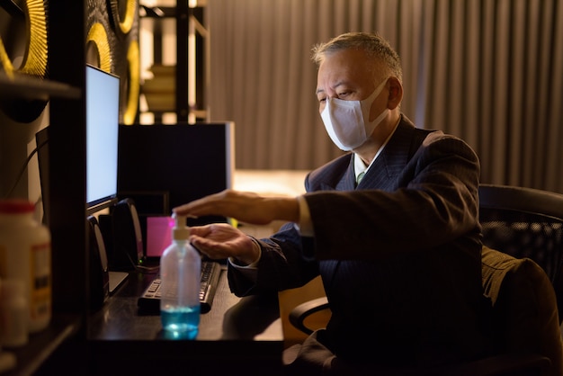 Mature Japanese businessman with mask using hand sanitizer while working overtime at home late at night