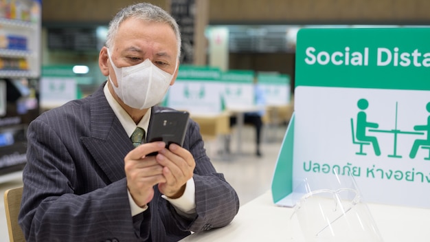 Mature Japanese businessman with mask using phone and sitting with distance at the food court