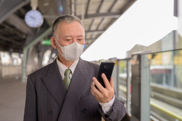 Mature Japanese businessman with mask using phone at the sky train station